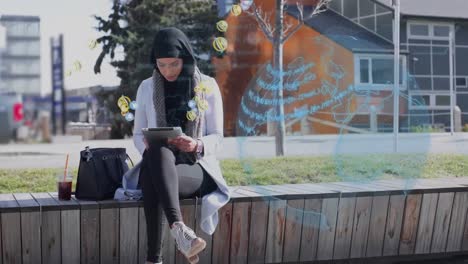 Globe-and-multiple-digital-icons-floating-over-woman-in-hijab-using-digital-tablet-on-the-street