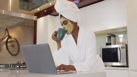 Afroamerikanische-Attraktive-Frau-Mit-Aufgesetzter-Gesichtsmaske,-Trinkt-Kaffee-Und-Benutzt-Laptop-In-Der-Küche