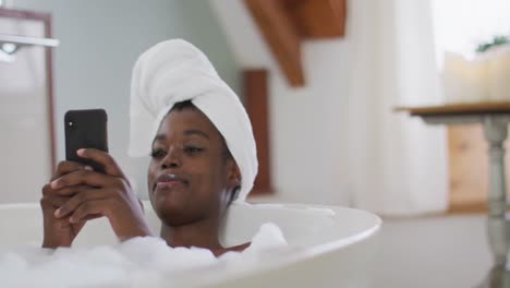 Lächelnde-Afroamerikanische-Attraktive-Frau,-Die-Sich-Im-Bad-Entspannt-Und-Im-Badezimmer-Ihr-Smartphone-Benutzt