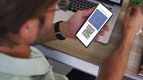 Man-at-desk-holding-smartphone-with-covid-vaccination-certificate,-eu-flag-and-qr-code-on-screen