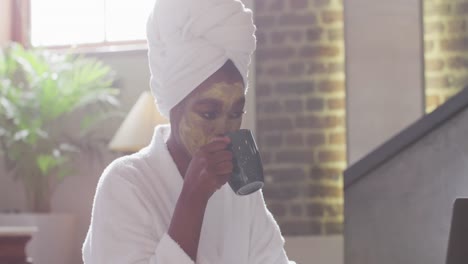 Afroamerikanische-Attraktive-Frau-Mit-Aufgesetzter-Gesichtsmaske,-Die-Zu-Hause-Kaffee-Trinkt-Und-Einen-Laptop-Benutzt