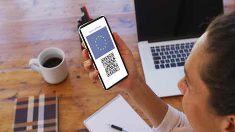 Woman-at-desk-holding-smartphone-with-covid-vaccination-certificate,-eu-flag-and-qr-code-on-screen