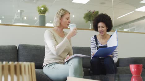 Diversas-Colegas-Femeninas-Sentadas-En-El-Sofá,-Tomando-Café,-Conversando-De-Negocios-En-La-Oficina