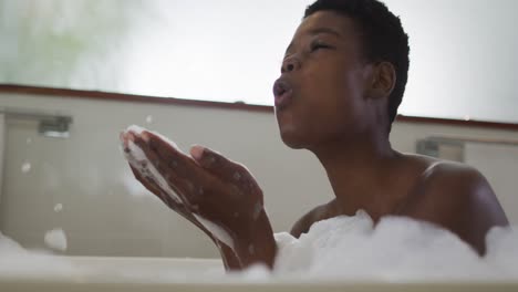 Smiling-african-american-attractive-woman-taking-bath-and-blowing-out-foam-in-bathroom
