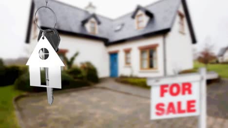 Animation-of-silver-house-key-fob-and-key,-hanging-in-front-of-blurred-house-with-for-sale-sign