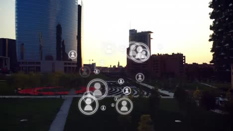 Animation-of-multiplying-people-icons-moving-over-modern-cityscape-at-sunset
