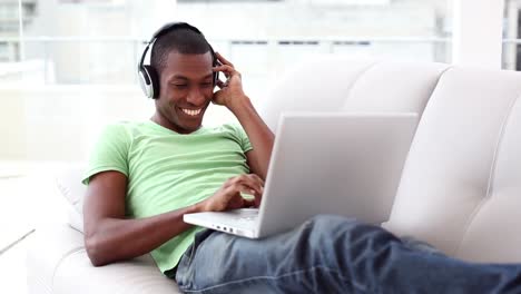 Smiling-man-lying-on-couch-listening-to-music-using-laptop