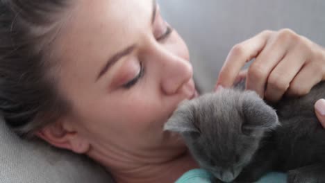 Smiling-woman-lying-on-couch-cuddling-tiny-kitten