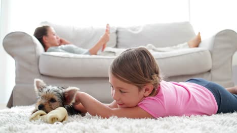 Little-girl-playing-with-puppy-chewing-bone-with-her-mother-reading-on-the-sofa