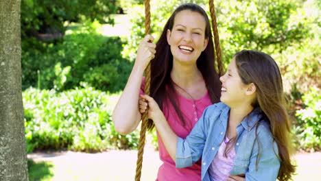 Mother-and-daughter-sitting-on-a-swing-in-the-park-waving-at-camera