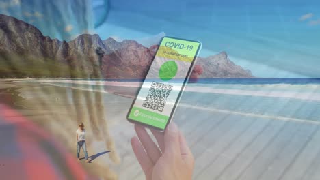 Woman-holding-a-smartphone-with-qr-code-on-screen-against-caucasian-woman-walking-on-the-beach