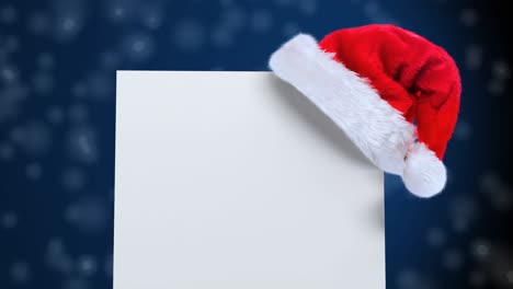 Weihnachtsmütze-über-Einem-Leeren-Plakat-Gegen-Schneeflocken,-Die-Auf-Blauen-Hintergrund-Fallen