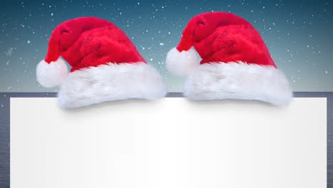 Two-santa-hats-over-a-blank-placard-against-snow-falling-on-blue-gradient-background