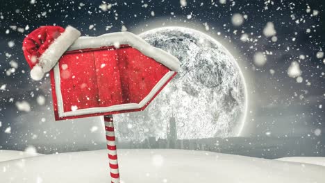 Snow-falling-and-santa-hat-over-red-wooden-sign-post-on-winter-landscape-against-moon-in-night-sky