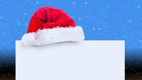 Animation-of-santa-hat-and-white-card-with-copy-space-over-snow-falling-on-blue-background