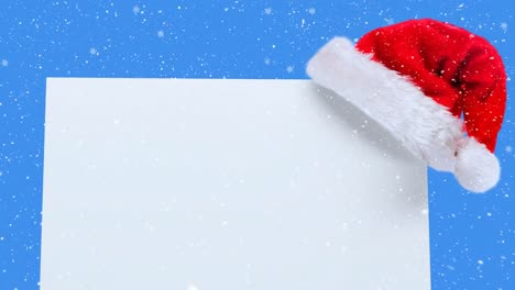 Animation-Von-Weihnachtsmütze-Und-Weißer-Karte-Mit-Kopierraum-über-Schnee,-Der-Auf-Blauen-Hintergrund-Fällt