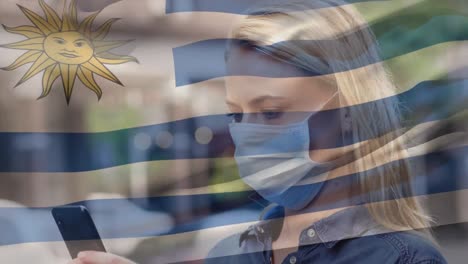 Animation-of-flag-of-uruguay-waving-over-caucasian-woman-wearing-face-mask-in-city-street