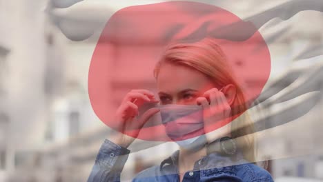 Animation-of-flag-of-japan-waving-over-caucasian-woman-wearing-face-mask-in-city-street