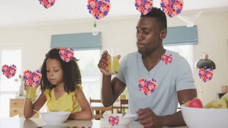 Animation-Von-Blumenherzen-über-Afroamerikanischem-Vater-Und-Tochter,-Die-Zu-Hause-Saft-Trinken