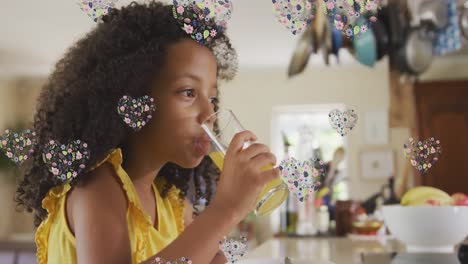 Animation-Von-Blumenherzen-über-Einem-Afroamerikanischen-Mädchen,-Das-Zu-Hause-Saft-Trinkt