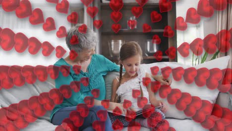 Animation-of-hearts-over-happy-caucasian-grandmother-and-granddaughter-reading-book-on-sofa