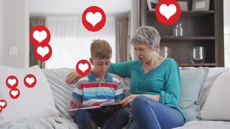 Animation-of-heart-icons-over-caucasian-grandmother-and-grandson-reading-book-in-livin-groom
