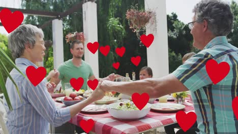 Animation-of-hearts-over-caucasian-family-praying-before-meal-in-garden