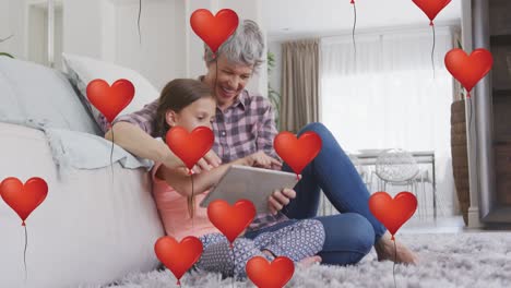 Animation-of-heart-balloons-over-happy-caucasian-grandmother-and-granddaughter-using-tablet-at-home