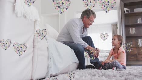 Animation-of-flower-hearts-over-grandfather-and-granddaughter-talking-in-bedroom