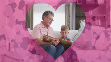 Animation-of-hearts-over-happy-caucasian-grandfather-and-grandson-reading-book-at-home