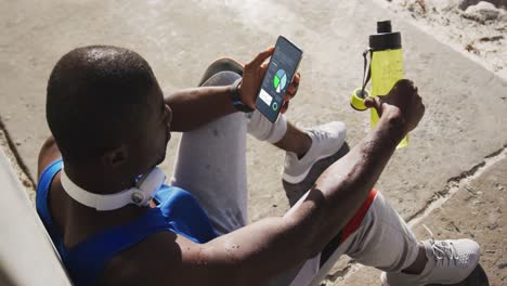 African-american-sportsman-sitting-at-pavement,-using-smartphone-with-statistics-on-screen