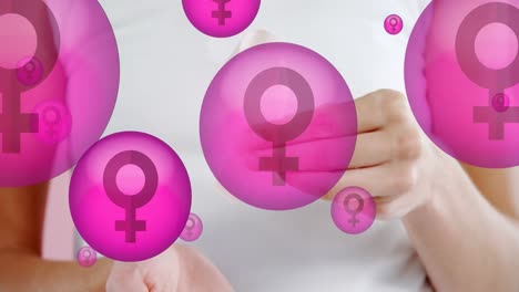 Animation-of-female-symbol-in-pink-bubbles-with-hands-of-woman-holding-breast-cancer-ribbon,-on-pink