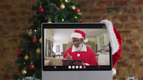 Hombre-Afroamericano-Mayor-Disfrazado-De-Papá-Noel-En-Videollamada-En-Computadora,-Con-árbol-De-Navidad
