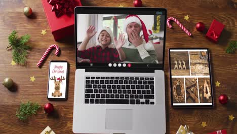 Father-and-son-wearing-santa-hats-on-video-call-on-laptop,-with-smartphone,-tablet-and-decorations