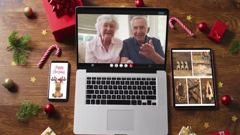 Caucasian-senior-couple-waving-on-video-call-on-laptop,-with-smartphone,-tablet-and-decorations