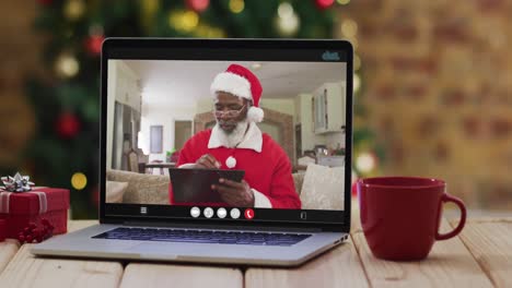 Hombre-Afroamericano-Mayor-Disfrazado-De-Santa-En-Videollamada-En-Computadora-Portátil,-Con-árbol-De-Navidad
