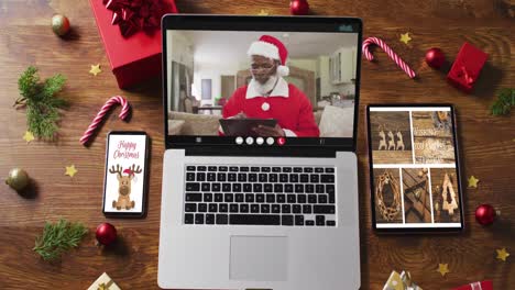 Senior-african-american-santa-on-video-call-on-laptop,-with-smartphone,-tablet-and-decorations