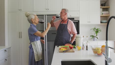 Caucasian-senior-couple-wearing-aprons-cooking-and-drinking-wine