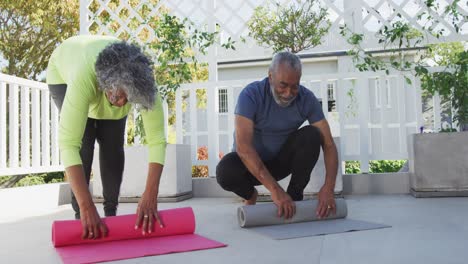 Glückliches-Afroamerikanisches-Seniorenpaar-Rollt-Yogamatten-Auf-Der-Terrasse