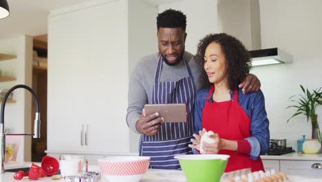Happy-african-american-couple-wearing-aprons,-using-tablet-and-baking-together