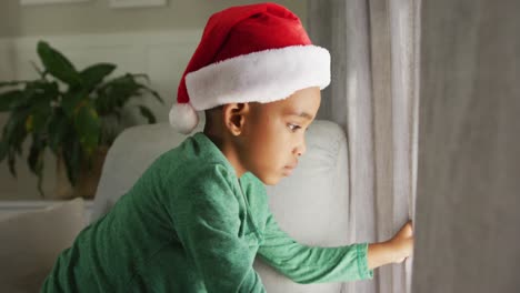 Sad-african-american-boy-wearing-santa-hat-looking-outside-window-at-christmas-time