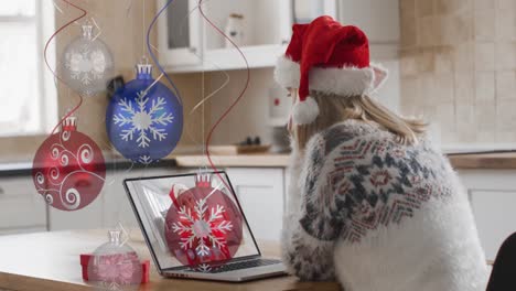 Animation-of-christmas-baubles-over-caucasian-woman-in-santa-hat-on-laptop-video-call-at-christmas