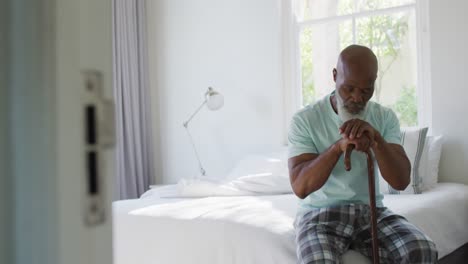 Thoughtful-senior-african-american-man-in-bedroom-holding-walking-cane