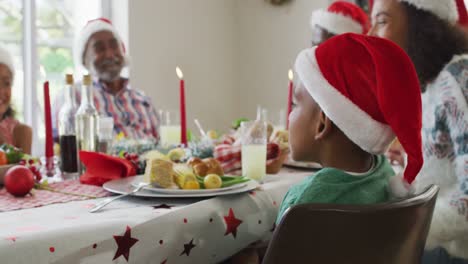 Portrait-of-smiling-african-american-boy-wearing-santa-hat-celebrating-holiday-meal-with-family