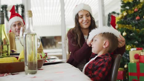 Glückliche-Kaukasische-Mutter,-Die-Ihrem-Sohn-Am-Weihnachtstisch-Eine-Weihnachtsmütze-Aufsetzt