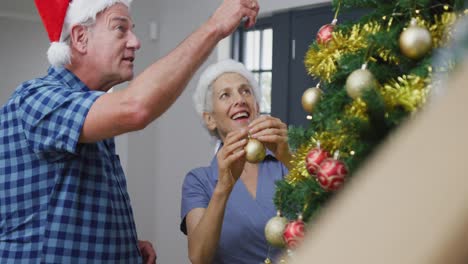 Happy-caucasian-senior-couple-wearing-santa-hats-decorating-christmas-tree