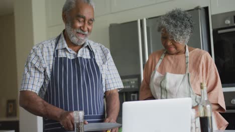Happy-african-american-senior-couple-cooking-together,-using-laptop
