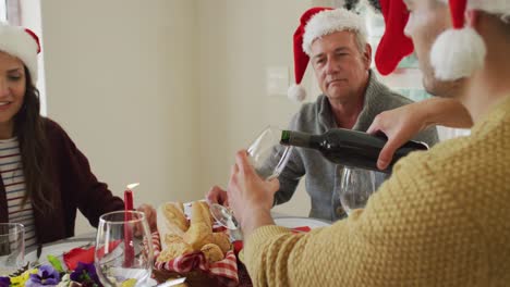 Glückliche-Kaukasische-Familie-Mit-Mehreren-Generationen,-Die-Weihnachtsmützen-Trägt,-Weihnachtsessen-Genießt-Und-Wein-Einschenkt
