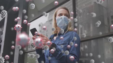 Animation-of-virus-cells-over-caucasian-female-wearing-face-mask,-using-smartphone