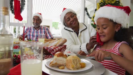 Fröhliches-Afroamerikanisches-Mädchen-Mit-Weihnachtsmütze-Feiert-Feiertagsessen-Mit-Großeltern
